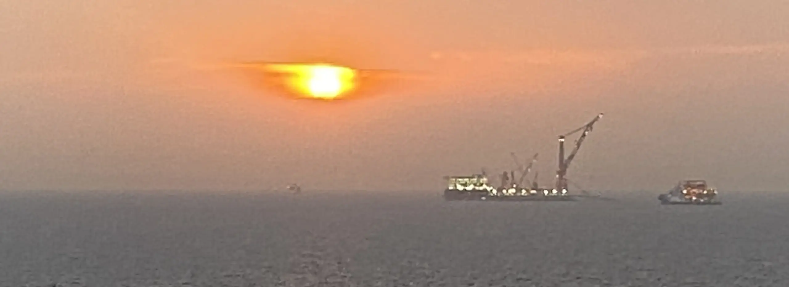 Offshore vessels and cranes silhouetted against a glowing orange sunset over calm sea waters.